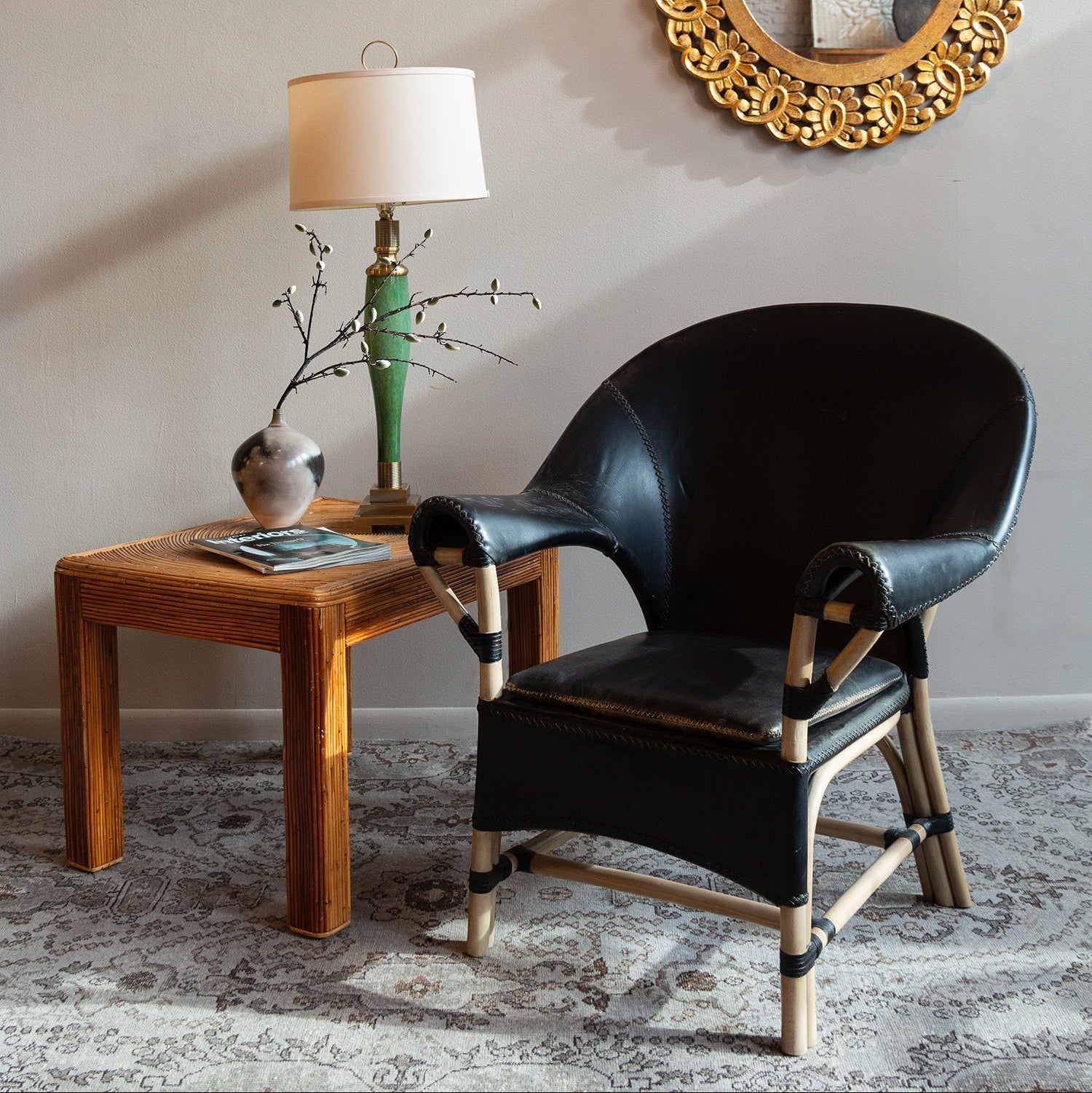Black leather chair with wooden legs next to a wooden side table with a lamp and decorative items, against a wall with a decorative mirror.